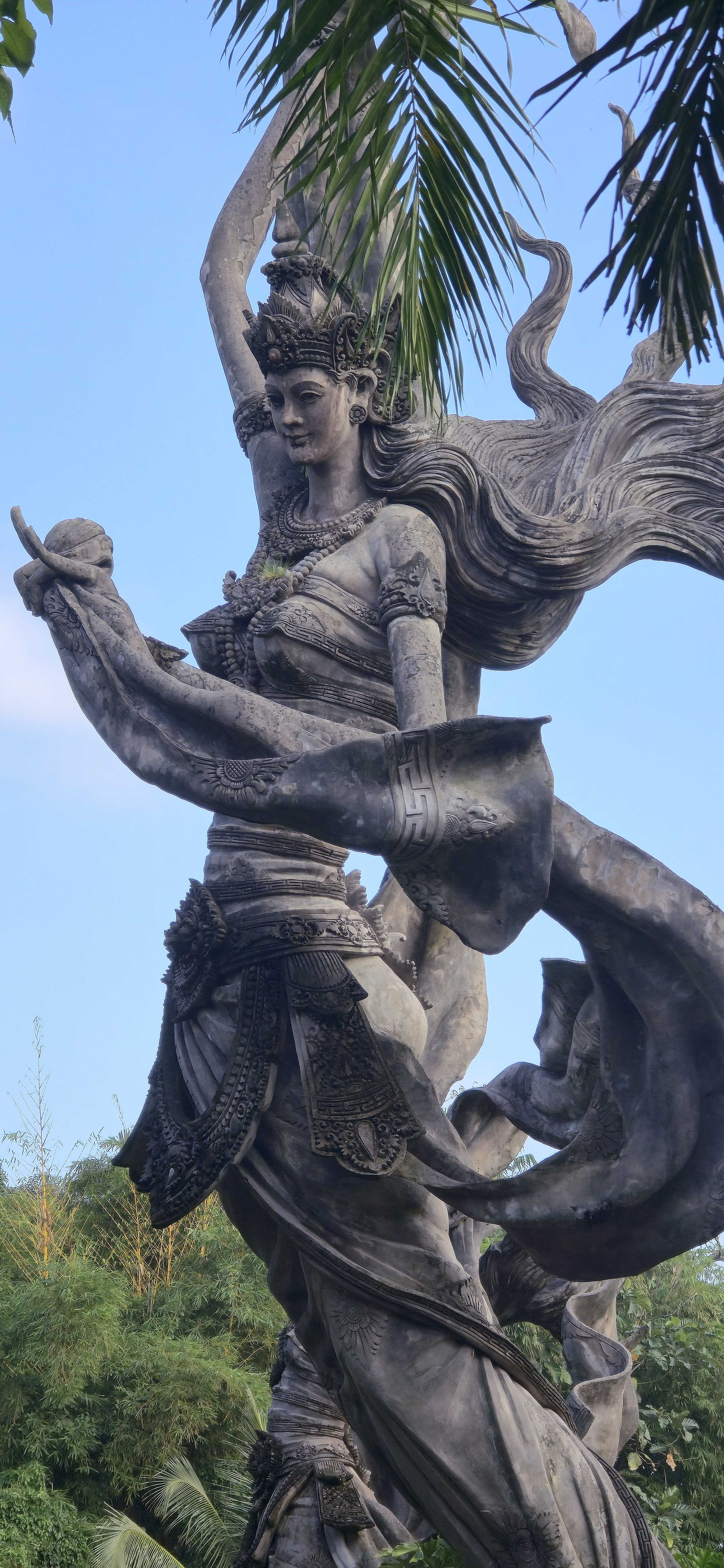 Stone statue of a woman with flowing hair holds a curved object. Intricate details, set against a clear blue sky and palm leaves.