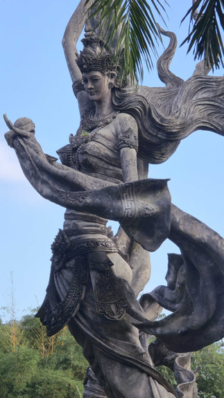 Stone statue of a woman with flowing hair holds a curved object. Intricate details, set against a clear blue sky and palm leaves.