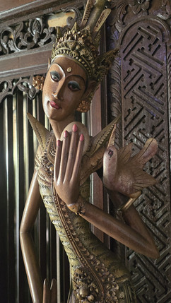 Ornate carved wooden statue of a woman with a crown and bird, posed beside a patterned wooden panel.