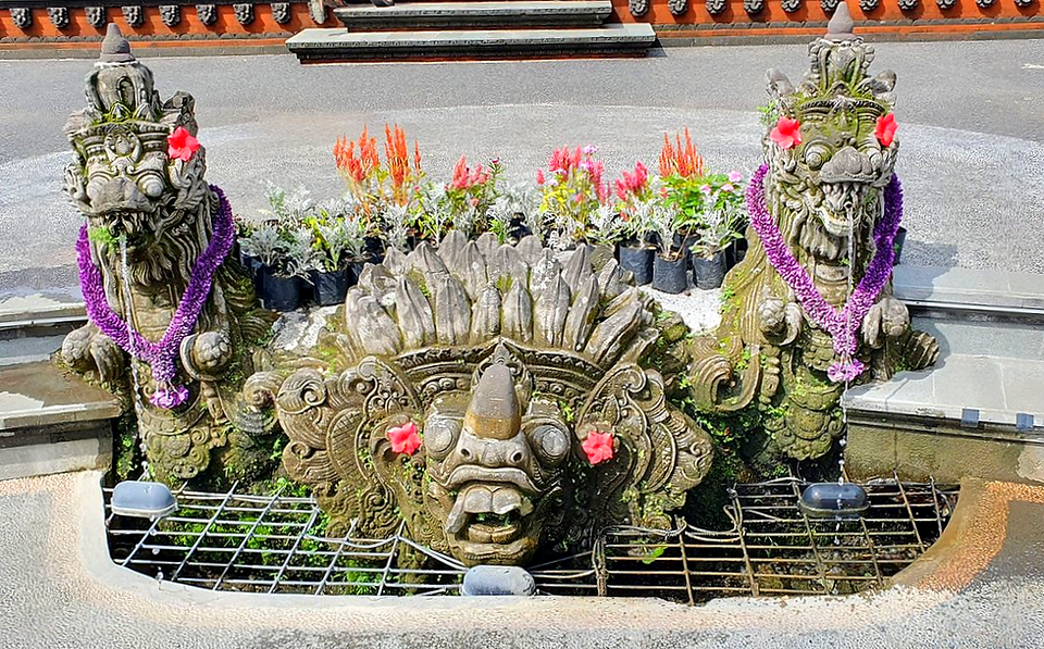 3 ornate stone statues with red flowers and purple garlands, surrounded by colourful potted flowers, spouting water in a courtyard.