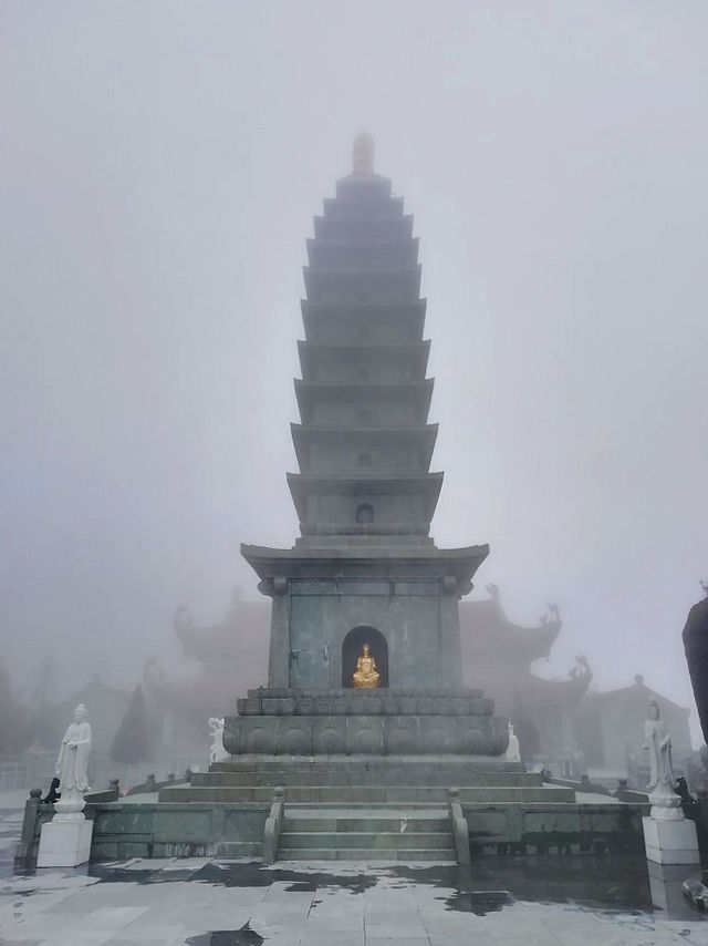 A misty temple scene with a tall stone pagoda and golden Buddha statue. Statues surround the base. Fog creates a serene and mystical mood.