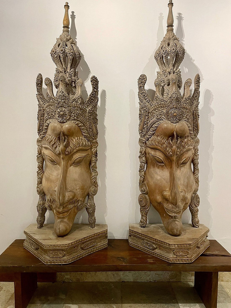 Two ornate carved face statues with crowns sit side by side on a wooden table against a plain white wall.
