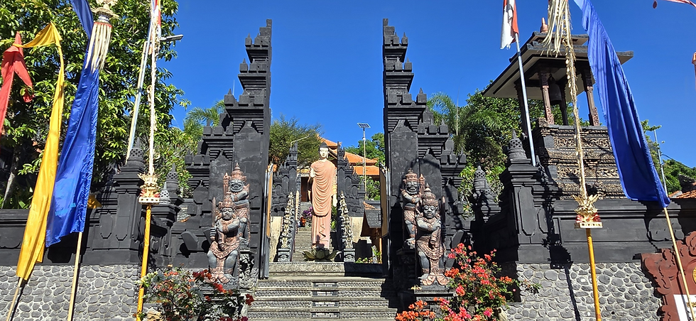 Balinese temple gate with black stone towers, guardian statues, and a Buddha statue framed by colorful flags under a bright blue sky.