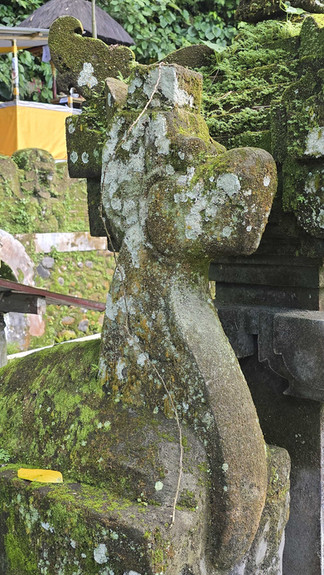 Moss-covered stone statue with dog-like features, set in lush greenery. A small yellow object lies on the statue, a temple structure in the background.