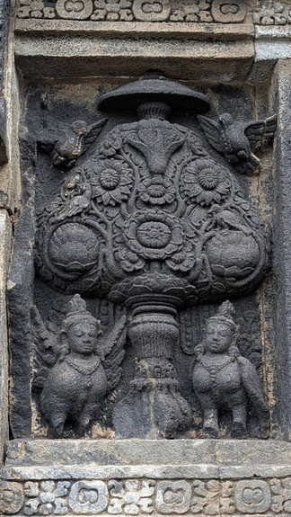 Ancient stone carving of two ornate figures, a stylized tree with floral details, and birds. The intricate grey and black relief exudes serenity.