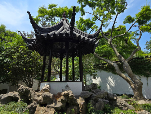 A traditional Chinese pavilion in a serene garden, surrounded by rocks and lush green trees, under a clear blue sky.