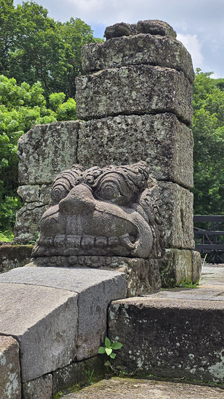 Stone sculpture of a creature's face on ancient steps, surrounded by lush green trees and a partly cloudy sky in the background.