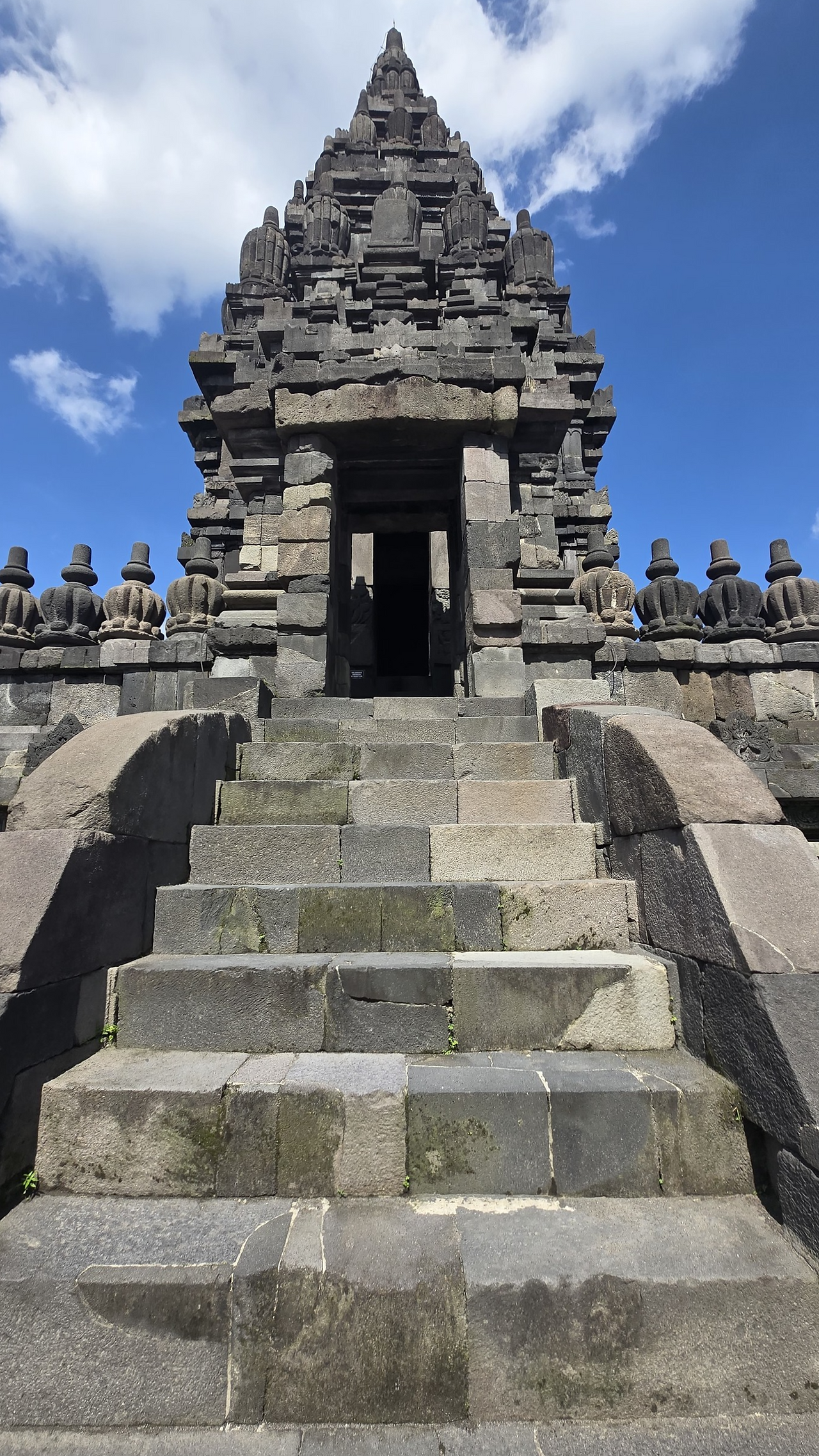 Ancient stone temple with intricate carvings under a clear blue sky. Steps lead to a dark entrance. Stone statues sit along the edge.