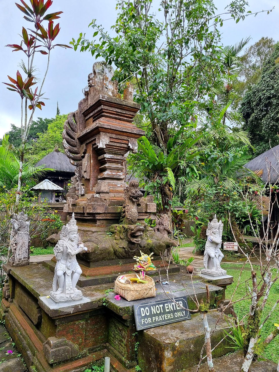 Stone temple with dragon carvings, flanked by statues and offerings. Sign reads "Do Not Enter, For Prayers Only." Lush green backdrop.