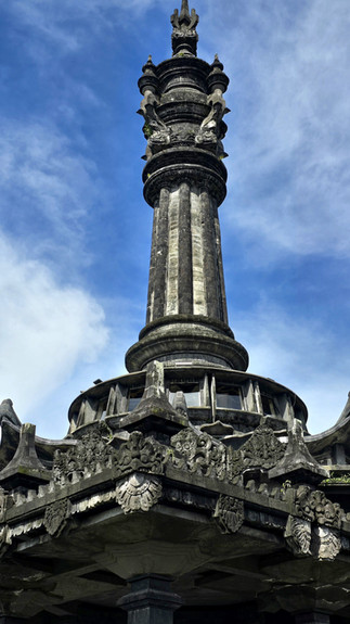 Tall stone monument with intricate carvings and spire, set against a blue sky. The structure has a circular base with ornate details.