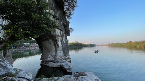A stone arch with greenery overlooks a calm river under a blue sky. A small boat with people sails in the distance. A serene scene.