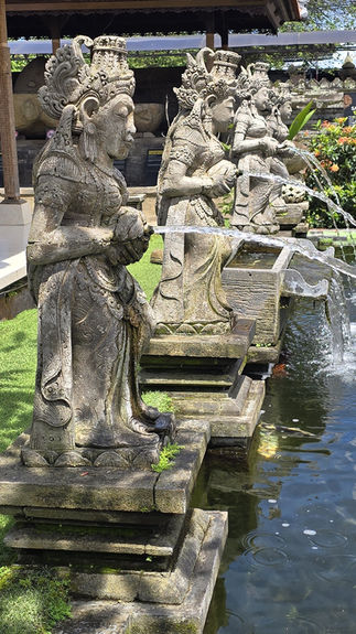 Stone statues in a courtyard fountain stream water into a pond. Background has lush plants, old stone structures and a red-tiled roof.