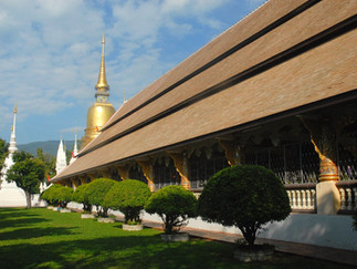 Buddhist temple with golden stupa, white pagodas, and lush green trees in the foreground. Blue sky and mountains in the background.