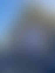 A tall standing Buddha draped in white, with both hands raised in a gesture of reassurance, set against the vibrant blue backdrop of the temple compound