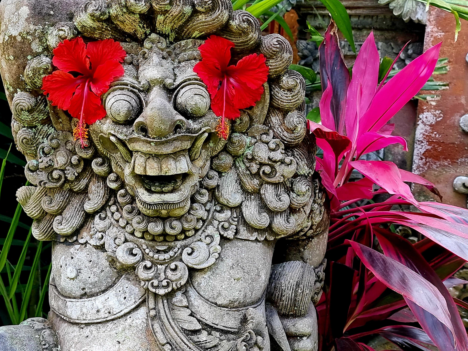 Stone statue with a joyful face adorned with red hibiscus flowers as eyes, surrounded by vibrant pink and green tropical leaves.
