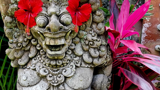 Stone statue with a joyful face adorned with red hibiscus flowers as eyes, surrounded by vibrant pink and green tropical leaves.