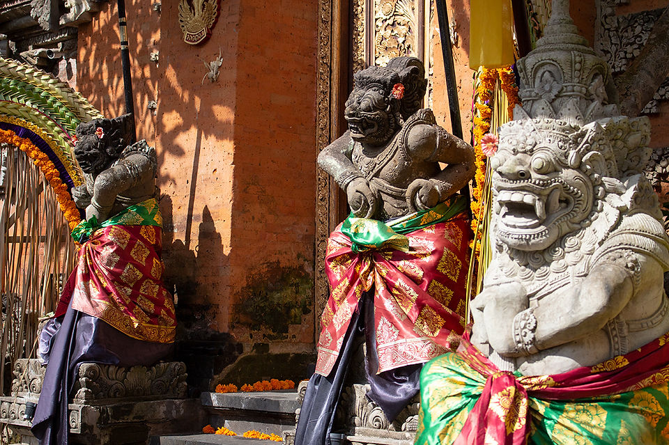 Stone statues with fierce expressions wear vibrant red and green cloths, adorned with flowers, against an ornately carved, sunlit wall.