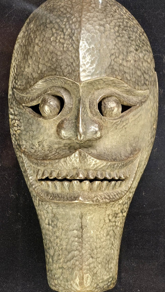 A textured metallic mask with a smiling face, prominent eyes, and mustache against a dark background. The mask has a weathered look.