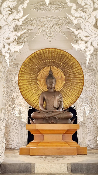 Sitting Buddha statue with a golden halo in an ornate, white temple setting. The detailed carvings add a serene and spiritual atmosphere.