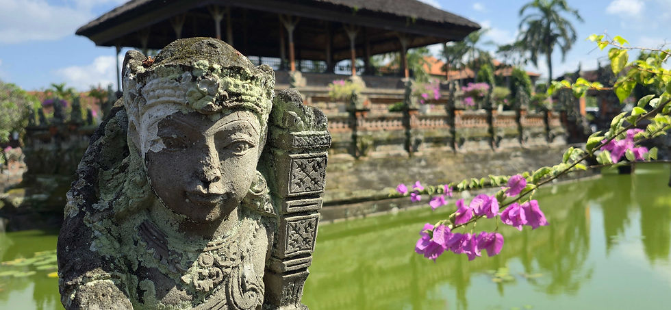 Stone statue with moss in foreground, pink flowers nearby. Background shows a thatched pavilion and green pond under a blue sky.
