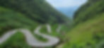Winding mountain road with green hills; three motorcyclists navigate sharp curves. Misty mountains in the distance under a cloudy sky.