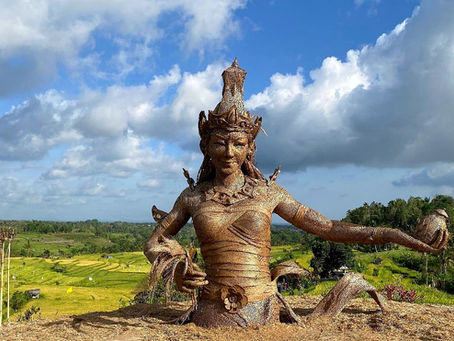 Bronze statue of a woman with ornate headpiece holds fruit, set against lush green rice paddies and blue sky with clouds. Tranquil mood.