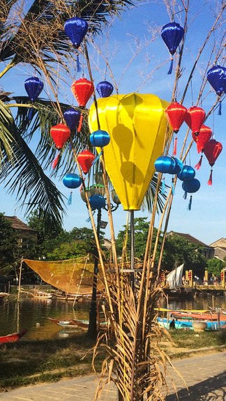 Colourful lanterns on bamboo poles by a river, with palm trees and boats in the background under a clear blue sky. Tranquil atmosphere.