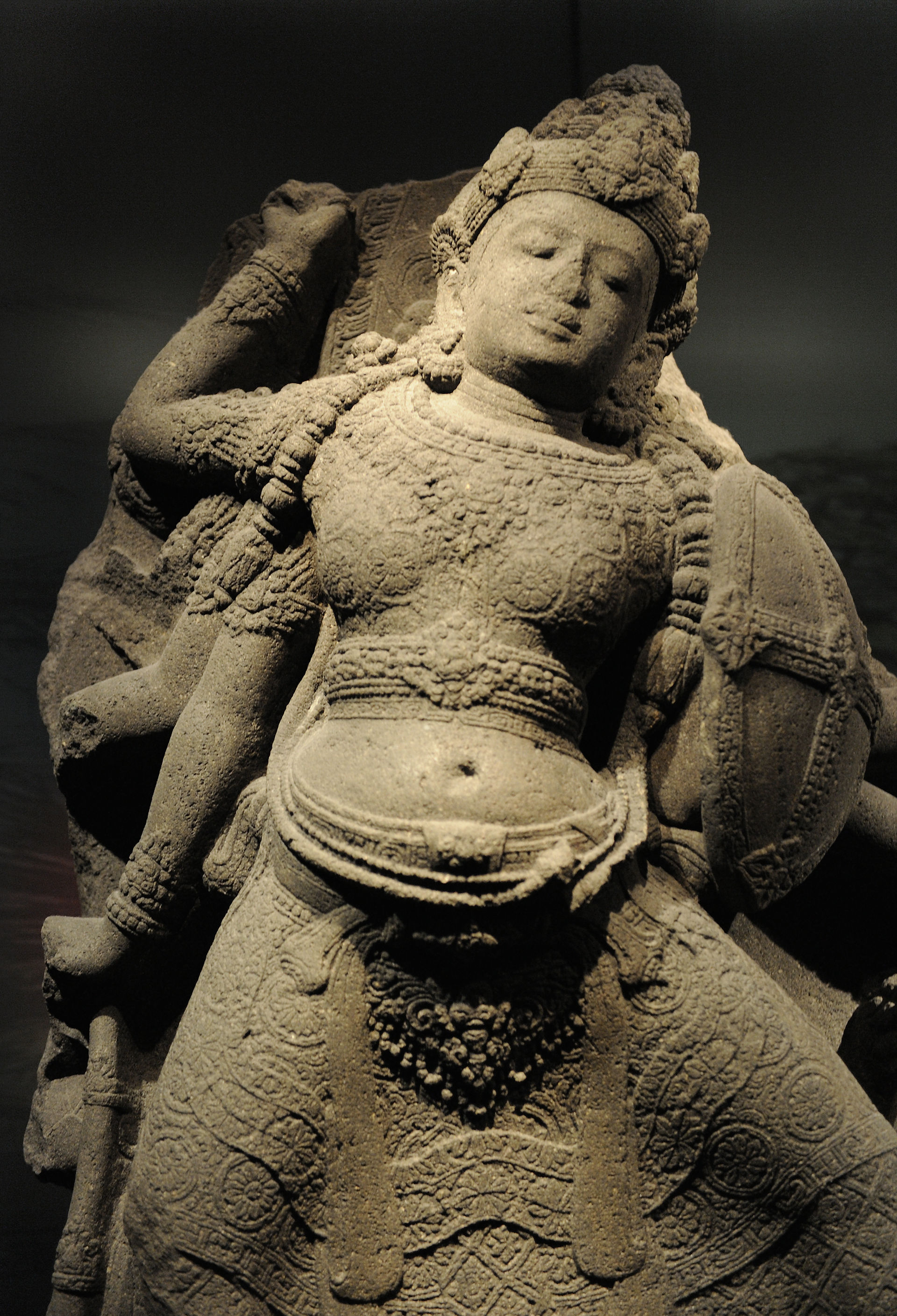 Ancient stone sculpture of a deity with intricate carvings, wearing ornate headdress. Soft lighting highlights textures in a museum setting.