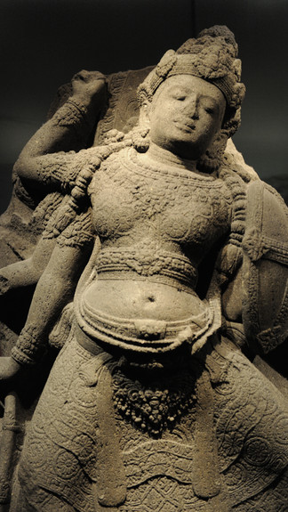 Ancient stone sculpture of a deity with intricate carvings, wearing ornate headdress. Soft lighting highlights textures in a museum setting.