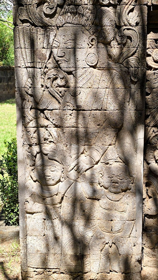 Sunlit stone relief of two ancient carved figures in a ruined wall, with shadows and ornate patterns outdoors.