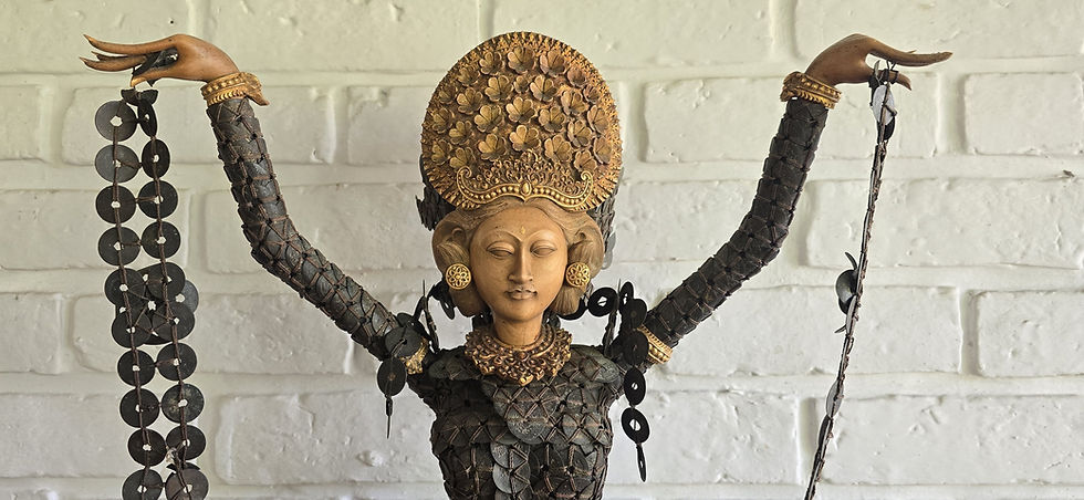 Decorative woman kepeng statue with raised arms, gold headdress and black jewelry, posed against a white brick wall.