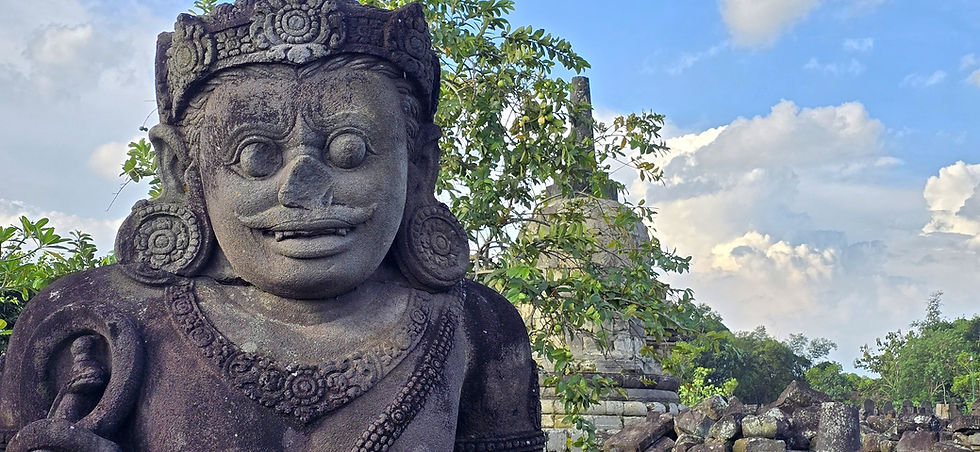 Stone statue with a carved face and intricate designs in a lush outdoor setting, cloudy sky in the background.