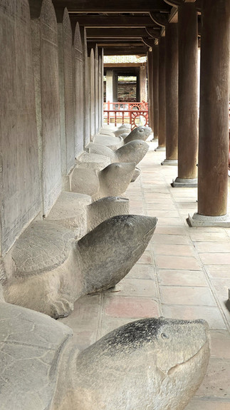 Stone turtles line a corridor in a historic setting, beneath wooden beams. The scene is serene, with muted earth tones and intricate carvings.
