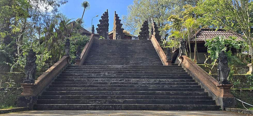 Wide stone staircase leads to a Balinese temple gate, flanked by carved statues and lush greenery under a bright blue sky.