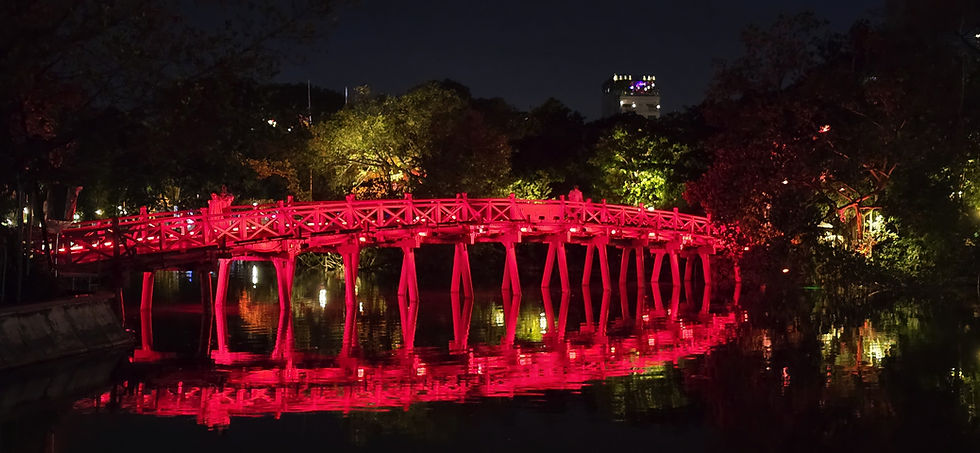 Red bridge at night over a calm river, reflecting vibrant red lights. Surrounded by lush trees and distant city lights in the background.