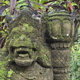 Moss-covered stone statue of a mythical creature with fierce expression in a lush, green forest setting. Bright foliage in the background.