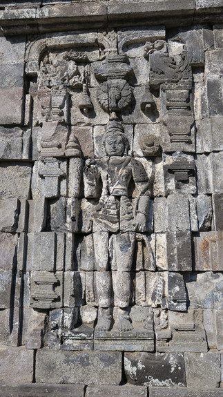 Ancient stone relief of a standing deity, adorned with intricate carvings, set against a weathered stone wall, evoking a historical atmosphere.