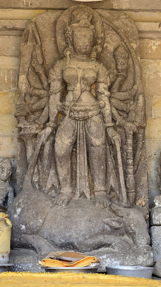 Ancient stone sculpture of Durga with multiple arms standing on a subdued animal. Gray tones dominate, set against a textured stone wall.