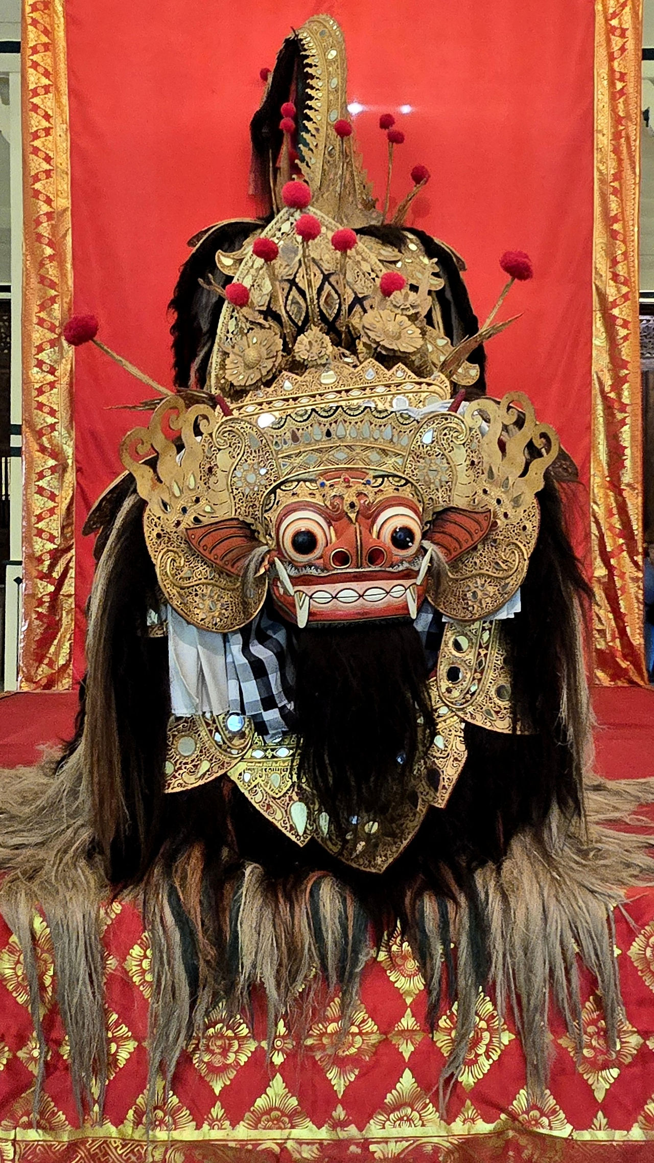 Intricate gold Barong mask with wide eyes and ornate details against a red and gold patterned backdrop