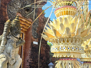 Stone guardian statue beside tall yellow ceremonial offerings in front of an ornate brick temple under blue sky.