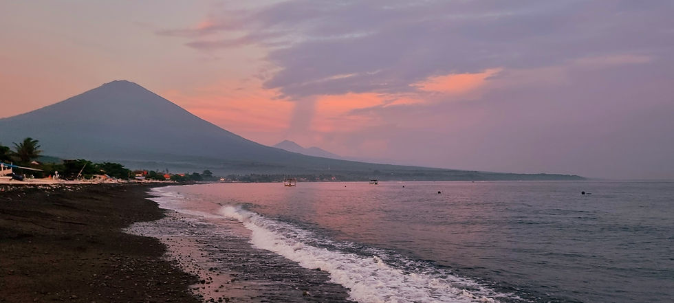 Pink sunset over a black-sand beach with gentle waves, a distant Mount Agung volcano and calm pastel sky.