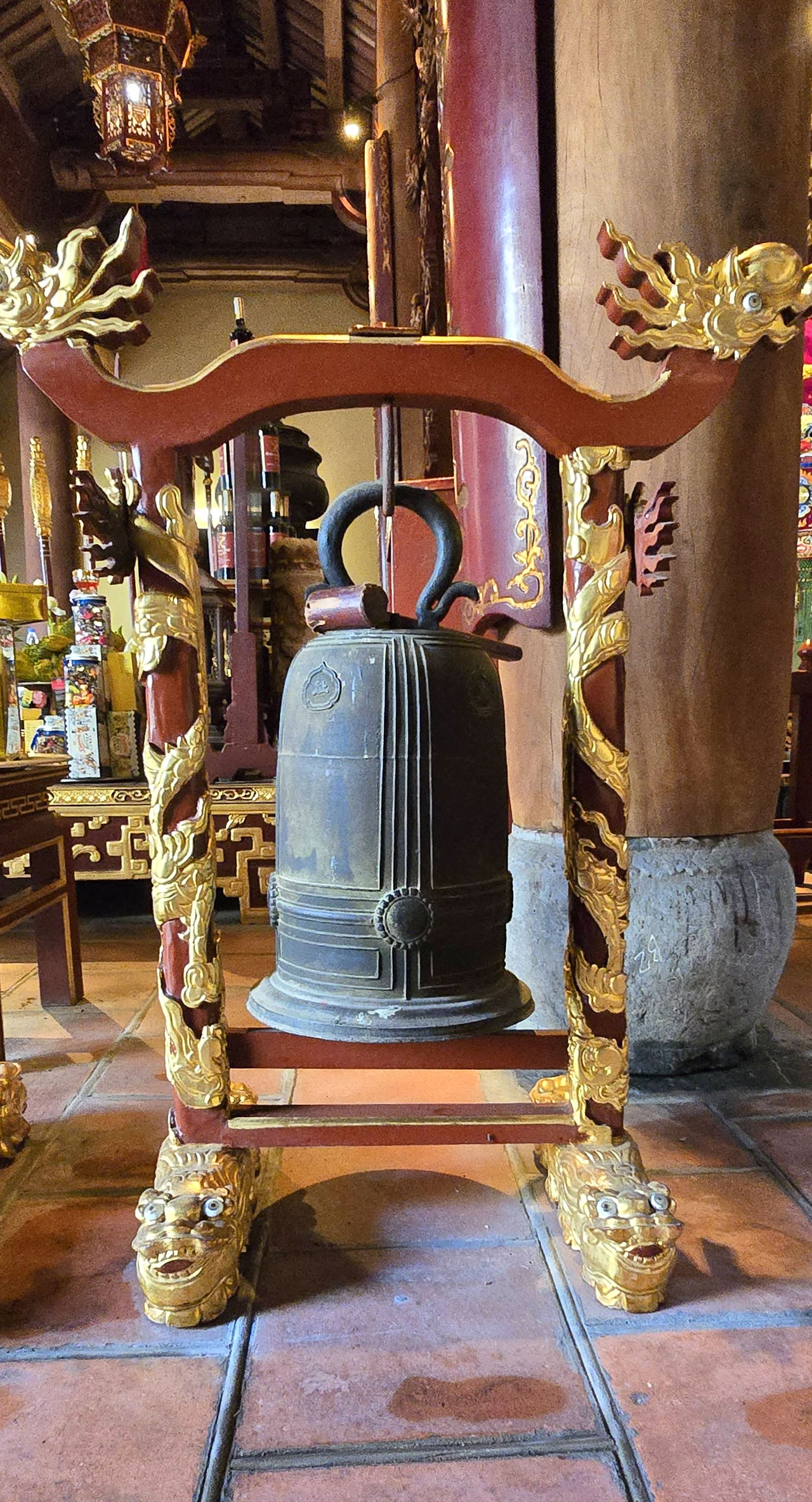 An ornate temple bell with dragon carvings and gold details, set in a wooden frame. The background shows intricate woodwork and tiled floors.