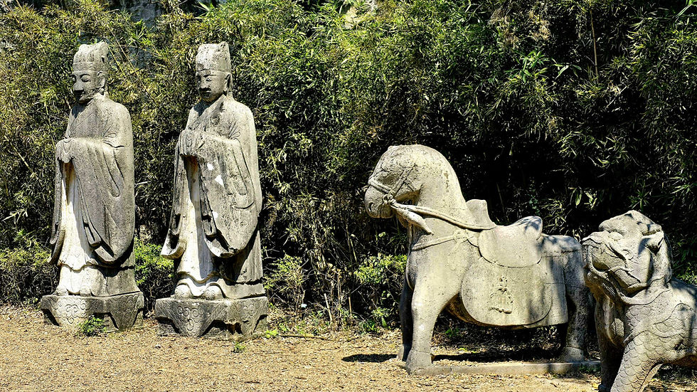 Stone statues of two robed figures, a horse and a lion stand in a lush garden. The scene is tranquil with greenery in the background.