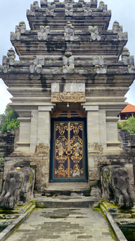Stone temple with ornate doors, elephant statues, and offerings on the ground. Moss-covered exterior, overcast sky, lush background.