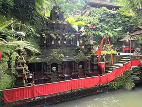 Lush green temple setting with moss-covered stone structures and red decorations, surrounded by ferns and trees, creating a serene atmosphere.