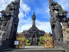 Ornate Balinese temple gate with stone statues and intricate golden gates. Towering spire in the background, set against a blue sky.