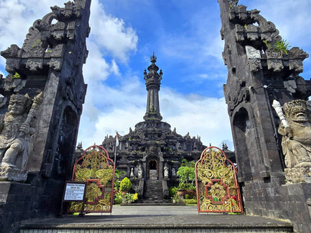 Ornate Balinese temple gate with stone statues and intricate golden gates. Towering spire in the background, set against a blue sky.