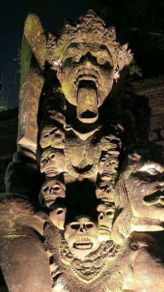 Stone statue at night with intricate carvings, including skulls and a fierce face showing a protruding tongue. Dimly lit, creating a dramatic mood.