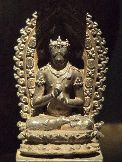Ancient bronze Buddha statue seated in meditation with ornate halo and headdress, set on a decorated pedestal against a dark background.
