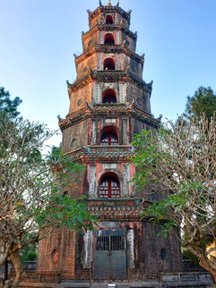 Tall pagoda with red and gray bricks, surrounded by trees under a clear blue sky. Intricate patterns and text on the structure. Peaceful mood.